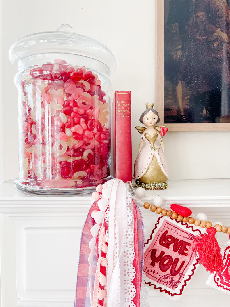 Glass jar filled with pink and red candy decorates a mantel beside a figurine and Valentine’s accents