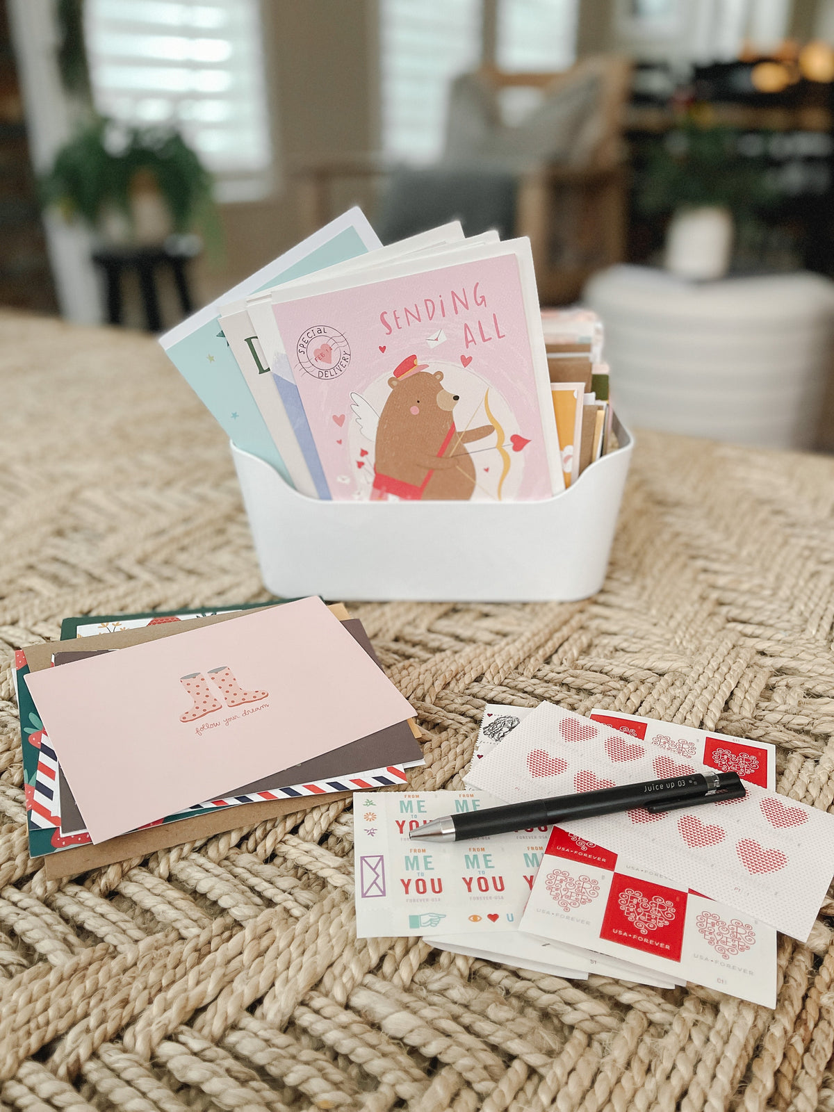 Stack of greeting cards and envelopes in a white organizer on a woven rug, stamps and a pen laid out for mailing, blurred living room background