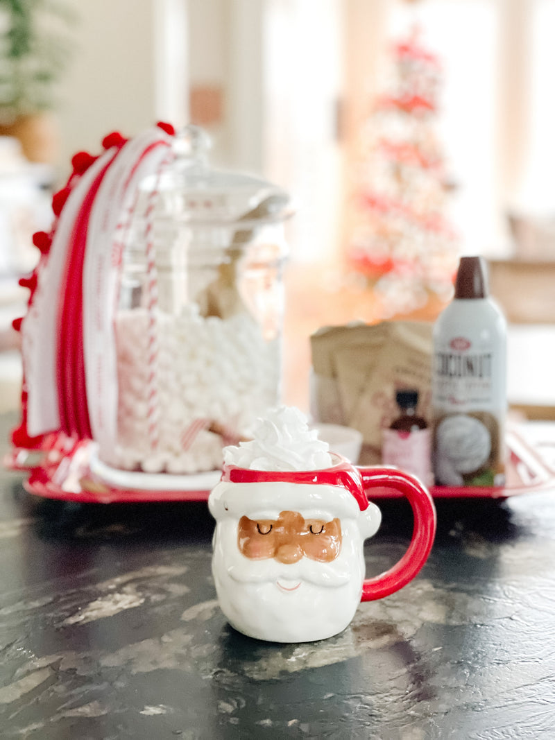 Santa mug with whipped cream on a counter, tray of marshmallows, cocoa packets and toppings blurred behind