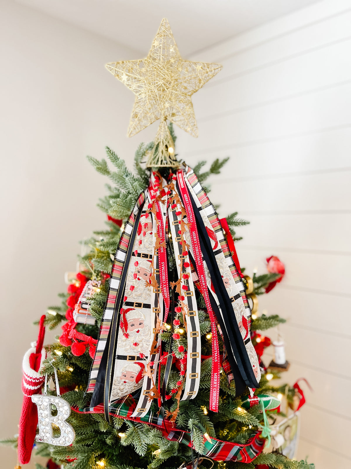 Christmas tree topper with gold star and long ribbons showing Santa faces and red and black stripes on white shiplap