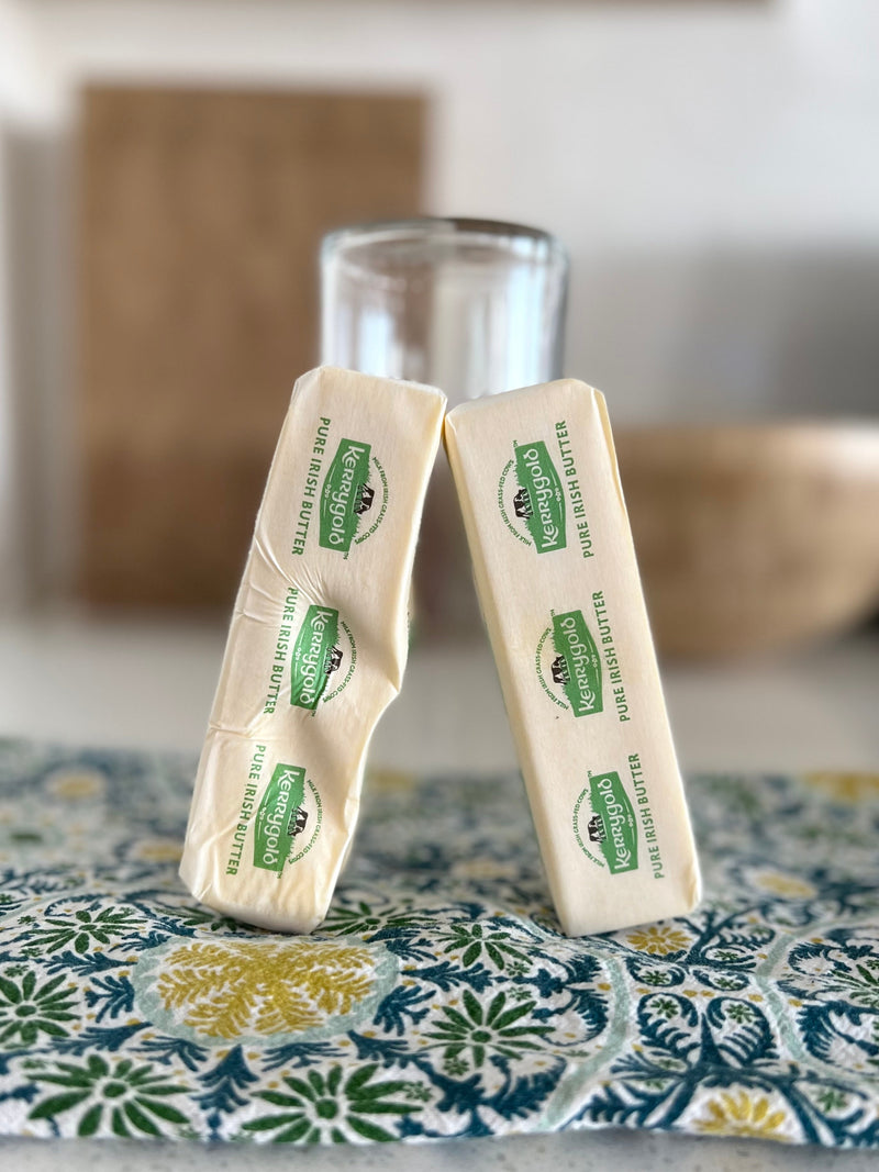Close-up of two wrapped Kerrygold Pure Irish Butter sticks leaning together on a patterned cloth with a blurred glass in the background