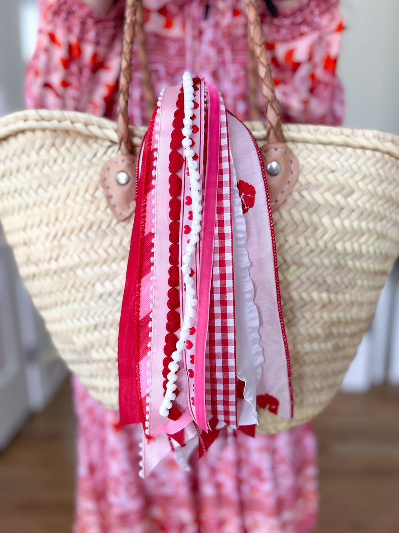 Pink and red Valentine-themed ribbons tied to a woven straw tote handle with a blurred pink patterned dress background