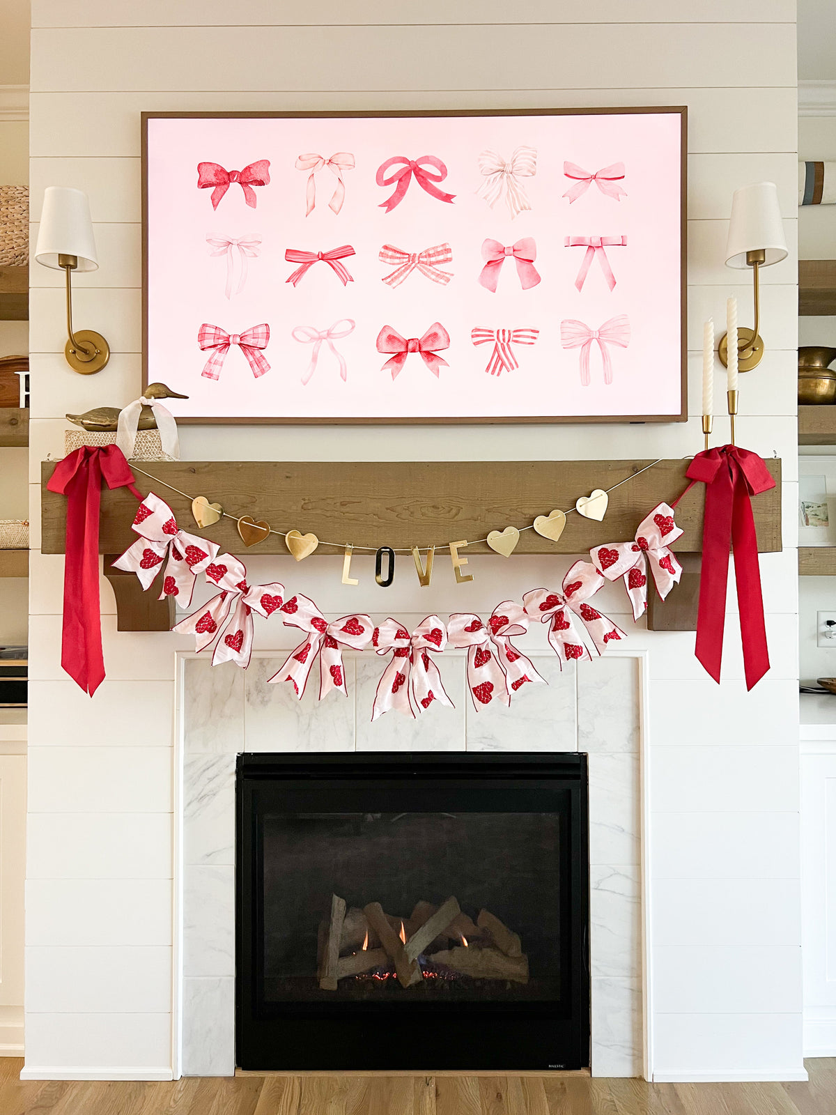 Fireplace mantel decorated for Valentine’s Day with framed pink bow artwork above, gold ‘LOVE’ garland, heart banner and red ribbon bows