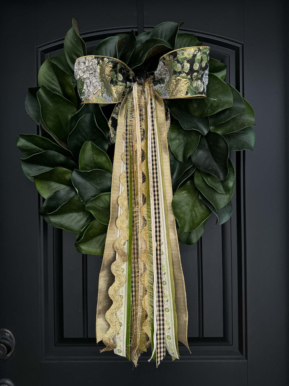 Close-up of magnolia leaf wreath showcasing a gold-and-green patterned bow and long mixed ribbons on a black front door