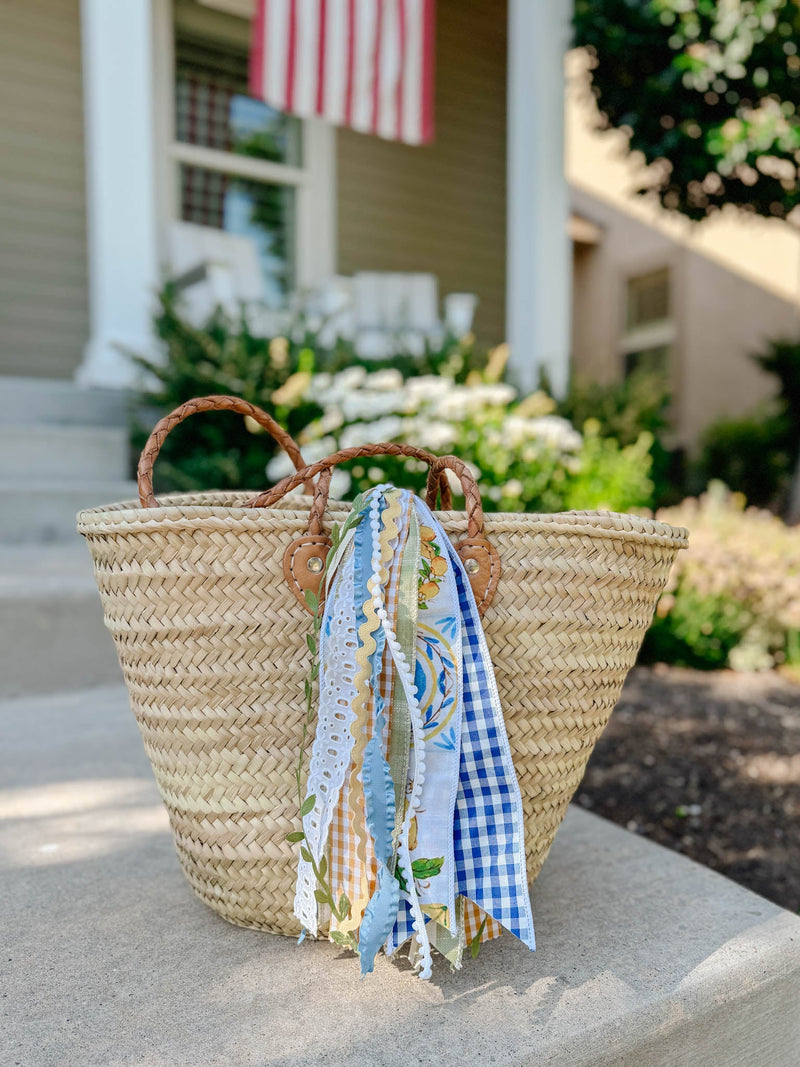 Woven straw tote with braided leather handles and ribbon tails on a front porch with an American flag and white flowers