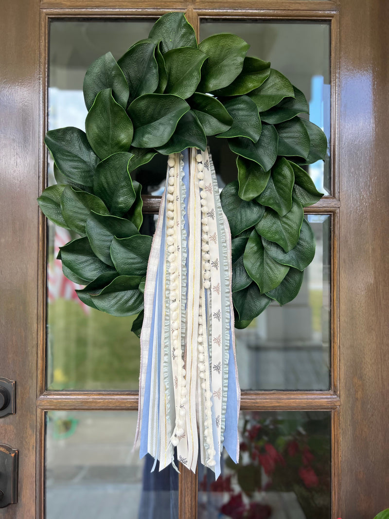 Front view of magnolia wreath with dark green leaves and long blue and cream ribbons with pom-pom trim on a wood door