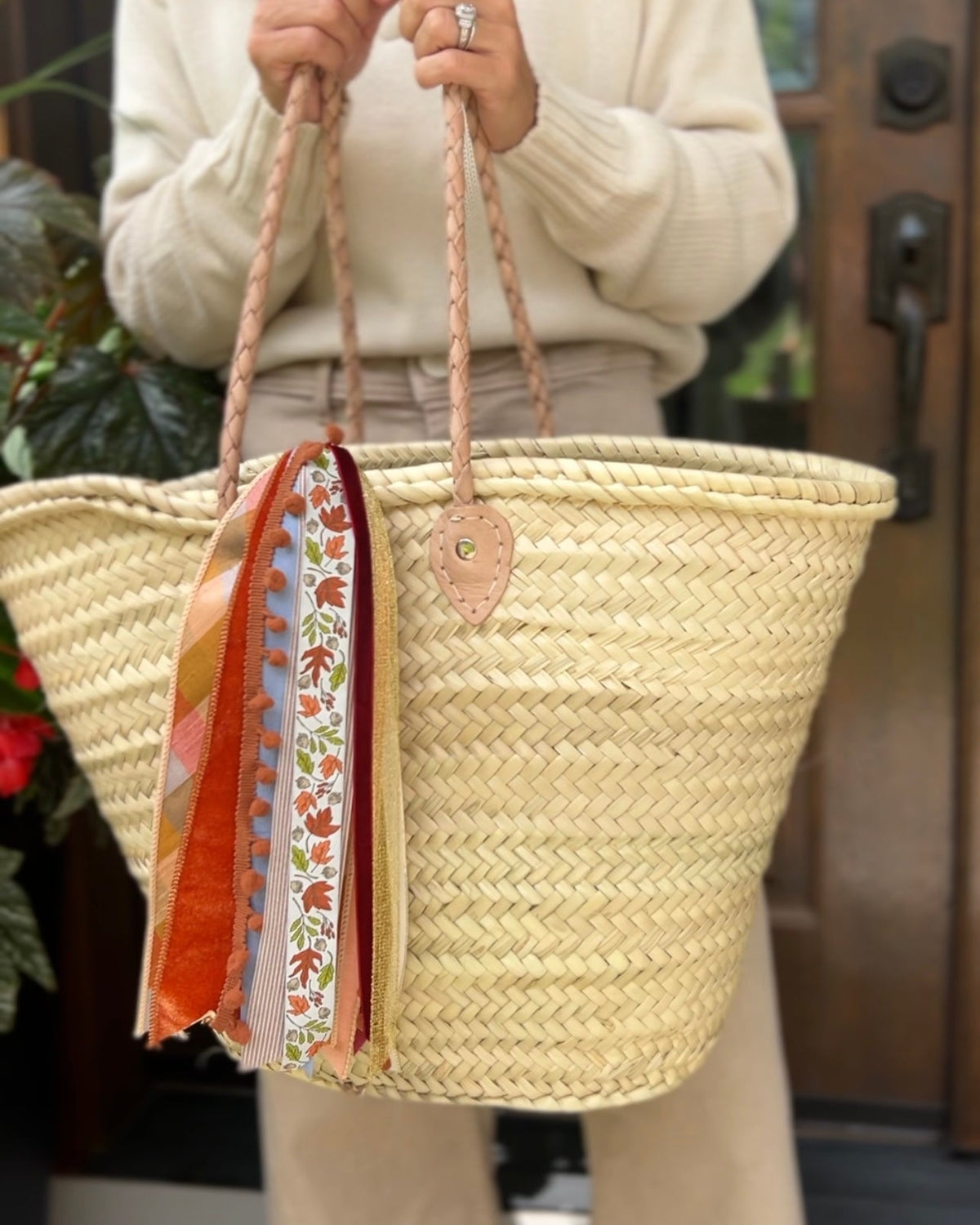 Person holding a woven straw basket with braided leather handles and fall ribbon tassels in front of a wooden front door