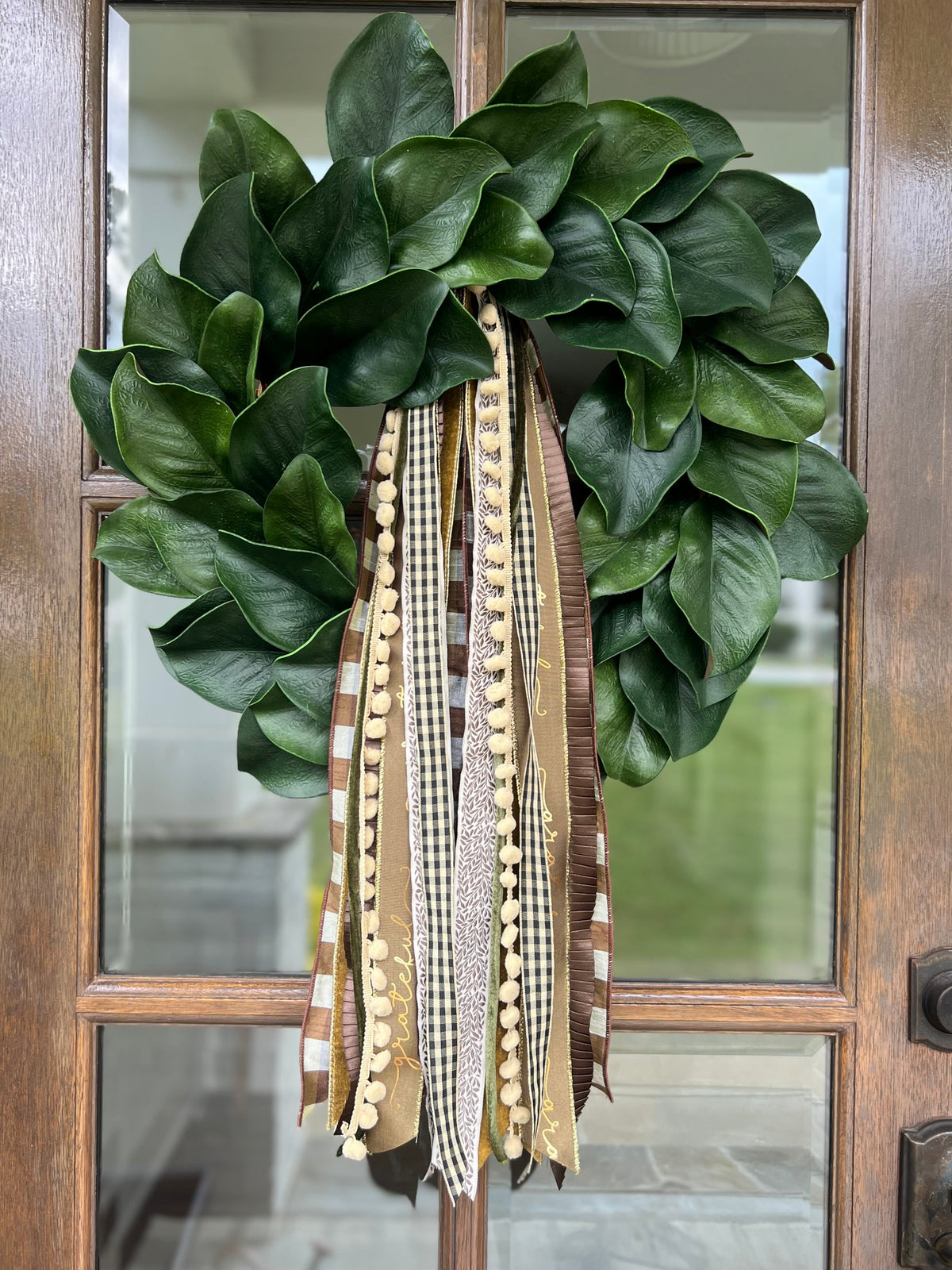 Close-up of magnolia wreath with glossy green leaves and layered brown-and-cream ribbon tails on a wooden door
