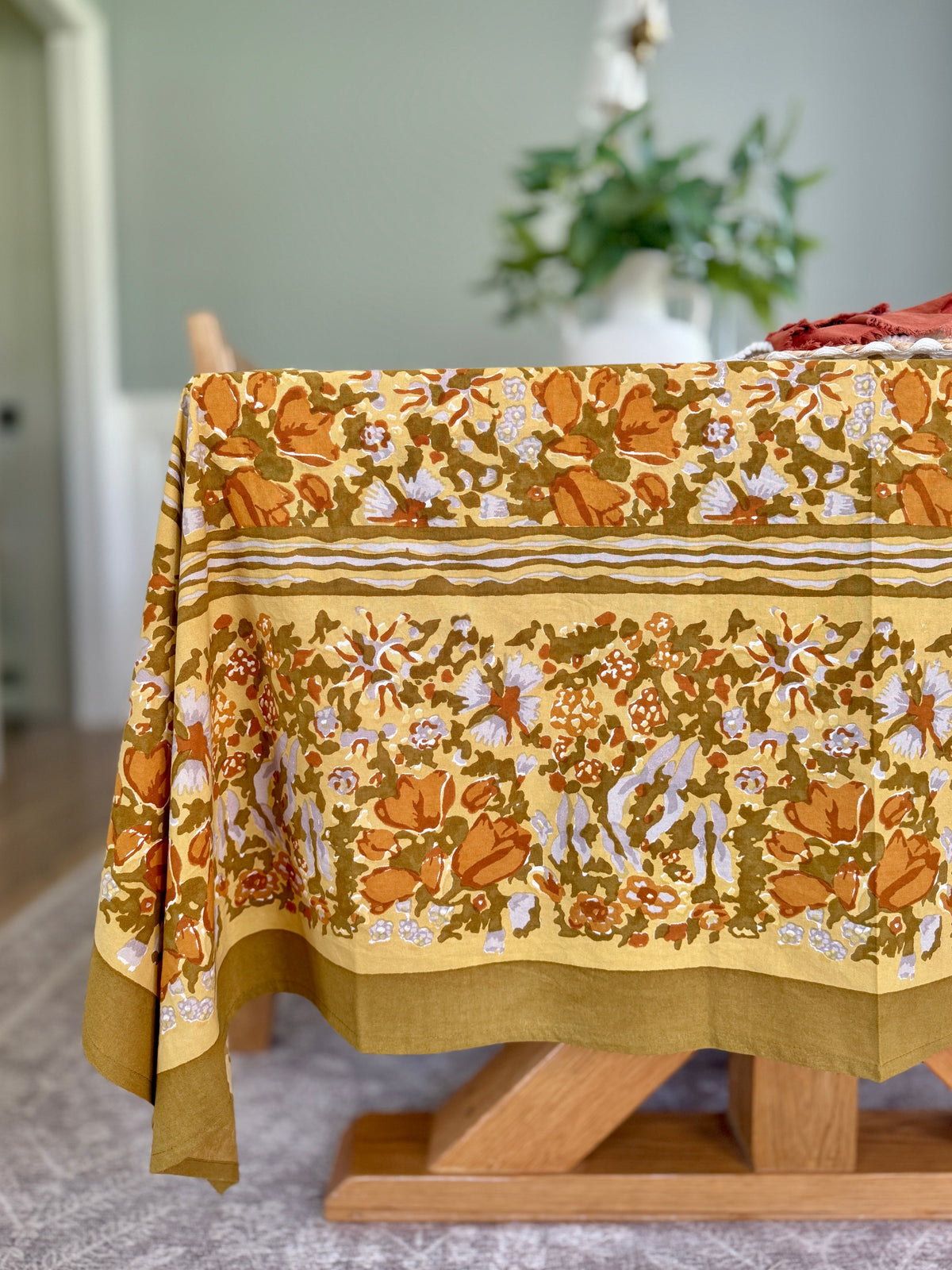Front edge of Golden Grove tablecloth showing orange floral print on mustard yellow fabric with wooden trestle table and blurred vase in background