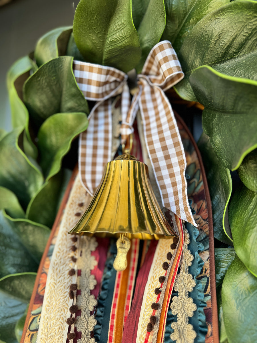 Brass bell hanging from brown-and-white gingham bow with layered decorative ribbons among green wreath leaves